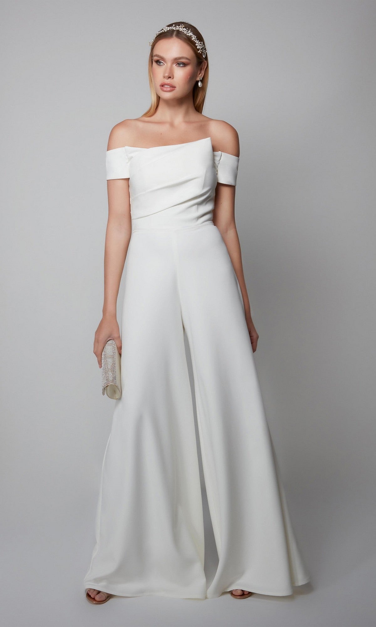 Woman in a white off-the-shoulder Long Formal Jumpsuit: Alyce 70014, holding a small clutch with straight hair and a decorative headband, stands against a plain light background.